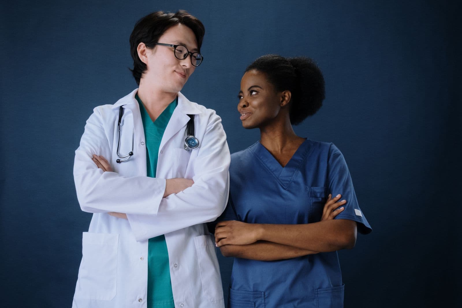Two people wearing a white coat and blue scrubs against a navy studio background